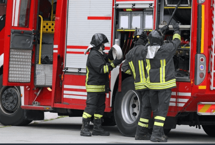 Firefighters in full gear, including helmets and protective clothing, are standing next to a red fire truck. One firefighter is holding a hose, while two others appear to be inspecting equipment on the truck. The scene appears to be part of an emergency response.